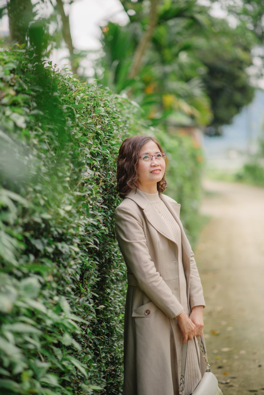 woman in a beige coat and dress standing by the hedge