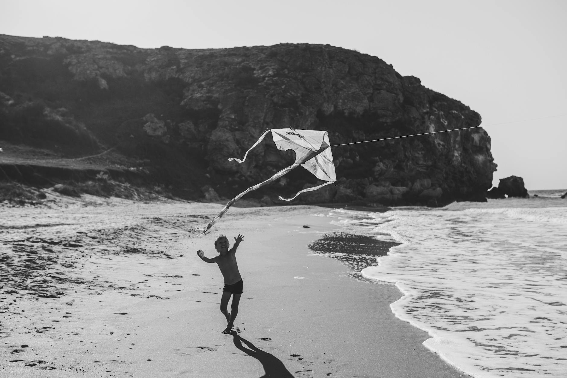 a boy in swimming trunks chasing a flying kite on shore